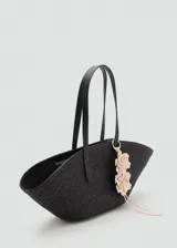 Bolso tipo capazo de fibra natural en color negro, con forma de media luna. Cuenta con dos asas largas de hombro en color negro y un detalle decorativo de flores en tonos claros colgando de una de las asas.