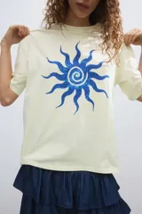 Remera oversized de algodón color amarillo claro con estampado frontal de sol azul. Presenta cuello redondo acanalado y mangas cortas con hombros caídos.