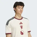 Camiseta de fútbol Adidas del Manchester United, correspondiente a la tercera equipación. Es de color blanco marfil con detalles en bordó y negro en el cuello, puños y hombros. Cuenta con el logotipo de Adidas, el escudo del club y un gráfico alineados verticalmente en el centro del pecho.