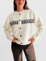 Cárdigan de punto color crema con diseño jacquard en la parte superior del pecho y cierre frontal de botones marrones. Presenta cuello redondo y puños acanalados.