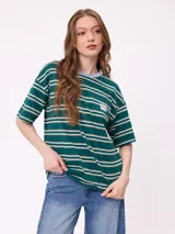 Remera de corte holgado con diseño de rayas horizontales en tonos beige, rosa y verde oliva. Presenta cuello redondo, mangas cortas y un bolsillo frontal pequeño con el logo de la marca.