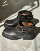 Bota corta de cuero color chocolate con suela track, cierre lateral y correa decorativa con hebilla metálica y tachas en el tobillo.