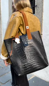 Bolso tote de cuero negro con textura de piel de cocodrilo, asas de cuero marrón y cierre con botón a presión.