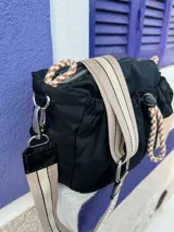 Cartera bandolera de nylon color negro con detalles en charol. Presenta un diseño fruncido con cordones decorativos combinados, grifa de marca en material sintético y correa regulable e intercambiable con logo estampado.