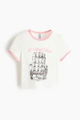 Remera corta entallada de algodón, color blanco, con ribetes rosados en el cuello y mangas, y estampa frontal de un edificio parisino con la inscripción "L'Hôtel Fleuri".