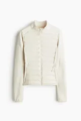 Campera híbrida color crema, con cuerpo acolchado y mangas de tejido elástico. Presenta cuello alto, cierre frontal y bolsillos laterales con cierre.