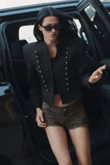 Blazer negro de corte entallado y manga larga con hombreras, cuello redondo y cierre frontal con gancho metálico. Presenta detalles de pasamanería bordada a tono y botones dorados en el delantero.