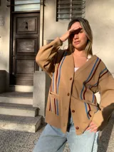 Cárdigan color beige de tejido de punto con rayas verticales azules y bordó. Tiene cuello en V, cierre frontal con botones marrones y bolsillos delanteros.