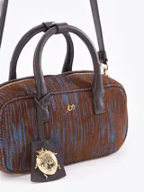 Cartera tipo baulito de cuero descarne vacuno con textura rayada en tonos marrón y azul. Presenta manijas cortas, cierre superior y un charm metálico dorado en forma de mariquita colgando de un tag de cuero. Incluye correa larga desmontable y regulable.