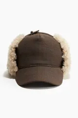 Gorra de tela con estampado a cuadros en tonos marrones y azules, con orejeras de sherpa suave que se abrochan con una trabilla ajustable en la parte superior. Forro de sherpa.