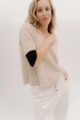 Buzo de punto en color beige con escote en V y mangas 3/4. Presenta un diseño holgado con tajos laterales y un detalle de contraste en color negro en la parte posterior de las mangas.