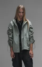 Campera rompevientos de diseño oversize, color verde, confeccionada en microfibra técnica ligera. Presenta capucha integrada, cierre frontal técnico y un sistema de frunces elásticos regulables a lo largo de las mangas que permiten ajustar el volumen de la prenda.