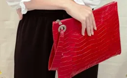 Sobre grande de cuero sintético rojo con textura tipo croco, cierre superior con cremallera y tira de cuero con argolla metálica.