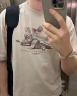 Remera de corte oversize color beige con efecto lavado, cuello redondo y mangas cortas. Presenta un estampado gráfico central en tonos oscuros con motivos de billetes y golosinas bajo la leyenda Kaboa Industries.