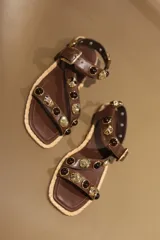 Sandalias bajas negras con tiras de cuero sintético, tachas plateadas y piedras rojas. Suela plana con borde de yute y cierre con hebilla.