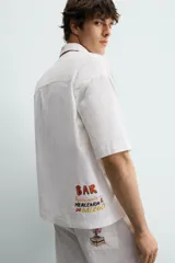 Camisa de corte relajado confeccionada en algodón, con cuello bowling y manga corta. Presenta un bolsillo en el pecho con vivo a contraste y bordados decorativos en el frente. Cierre frontal mediante botones.