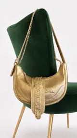 Cartera tipo bandolera de cuero color oro, de formato curvado, con una tira ancha central decorada con tachas y hebilla, cierre superior y correa mixta de cadena y cuero.