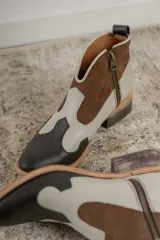 Botinetas estilo western de caña baja, confeccionadas en una combinación de cuero liso y gamuza en tonos marrón oscuro, marrón medio y crema. Presentan cierre lateral, taco bajo cuadrado y diseño con recortes característicos del estilo cowboy.
