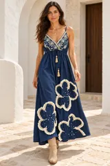 Vestido largo de denim azul oscuro con tirantes finos, escote en V y corte holgado. Presenta bordados ornamentales en el pecho y grandes apliques florales con flecos y perlas en la falda, además de un cordón ajustable en la cintura con borlas colgantes.