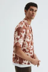 Camisa de manga corta con cuello abierto, tapeta de botones y bajo recto. Presenta un diseño estampado con motivos florales en tonos tierra y blanco.