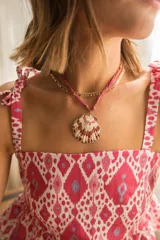 Collar compuesto por una cadena fina dorada con pequeños discos y una cinta de gasa teñida en tono rosado, que sostiene un dije central en forma de concha marina con bordes dorados.