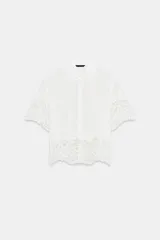 Camisa blanca de manga corta y cuello solapa, con detalles de bordados perforados y encaje en el borde inferior y las mangas. Cuenta con cierre frontal de botones.