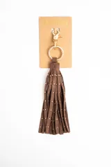 Charm para bolso compuesto por flecos de cuero sintético color marrón, decorados con pequeñas tachas metálicas doradas. Incluye un aro y un cierre de mosquetón metálico en tono dorado.
