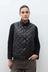 Chaleco capitoneado reversible con cuello alto, cierre frontal con botones a presión y bolsillos delanteros. Presenta un diseño bicolor en tonos verde oliva y beige con detalles en negro.