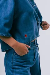 Camisa de denim azul de corte cropped, manga corta ancha, cuello camisero, cierre frontal de botones y un bolsillo aplicado en el pecho.