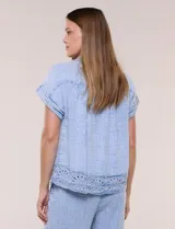 Blusa de algodón celeste con textura tramada tipo denim lavado, cuello solapa abierto en V, botones frontales y detalle de puntilla tejida al crochet en el ruedo.