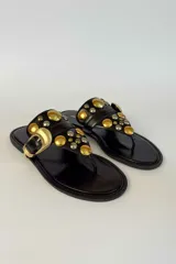 Sandalia plana de gamuza marrón, estilo ojota, con apliques metálicos dorados y hebilla decorativa.