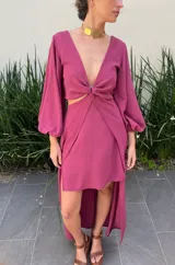 Vestido largo color bordó con escote en V profundo, detalle de nudo central y aberturas laterales en la cintura. Presenta mangas largas abullonadas con puños elásticos y falda con tajo frontal pronunciado.