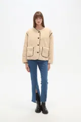 Campera de poliéster con textura de corderito en color beige, con cuello a la base y cierre frontal de botones. Presenta ribetes en contraste de color marrón oscuro en el cuello, la cartera, los hombros y en sus dos bolsillos frontales tipo parche.