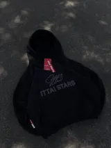 Hoodie de color negro con diseño de estampa frontal que combina una tipografía manuscrita con el texto ITTAI STARS en letras mayúsculas. Presenta un corte holgado, capucha amplia y bolsillo tipo canguro.