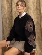 Campera de jogging negra con cierre frontal, cuello con detalle de volados y bordados florales en las mangas. Presenta un corte holgado y puños elásticos.