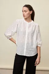 Camisa blanca de textura liviana con detalle de lunares en relieve, cuello mao y cierre frontal con botones. Cuenta con mangas 3/4 con puños elastizados y frunces en los hombros.