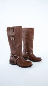 Bota de caña alta confeccionada en cuero color suela, con diseño de estilo western. Presenta tres hebillas metálicas en la parte superior de la caña y un estribo con argolla metálica en el tobillo. Cuenta con taco cuadrado de altura media.