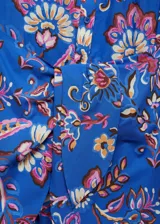 Vestido midi de algodón con estampado floral en tonos azules, rosas y crema. Presenta escote en pico, cierre frontal con botones y un lazo decorativo en la cintura que crea un efecto de nudo. Tiene mangas largas con puños abotonados y un diseño de corte recto.