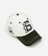 Gorra de béisbol de seis paneles, color blanco con visera curva en contraste de color verde militar. Presenta un bordado frontal de Snoopy junto a un banderín y el número cinco, y un bordado lateral con el texto 'Peanuts Athletic Department'.
