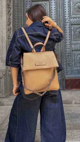 Mochila cartera color beige con textura granulada, diseño con tapa frontal y placa metálica con logo. Cuenta con correas ajustables y asa superior.