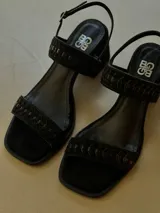 Sandalia de cuero negro con taco medio, punta cuadrada y tiras trenzadas. Cuenta con cierre de hebilla en el talón.
