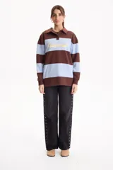 Buzo deportivo sin friza a rayas anchas en tonos blanco y tostado, con cuello polo, escote en V y estampa frontal con la inscripción "California est.1969" en color fucsia. Presenta un corte semi oversize y mangas largas.