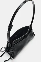 Bolso de hombro de material efecto acharolado negro, con doble compartimento exterior, cierre de cremallera y detalles de lazos en los laterales.