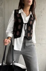 Chaleco negro de gamuza con bordados florales en tonos marrones. Presenta un cierre frontal con lazo y detalles de tachas metálicas en los bordes.