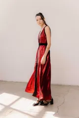 Vestido largo rojo brillante con escote en V profundo, espalda abierta baja y breteles anchos. Presenta detalles en terciopelo negro que enmarcan el escote, recorren el frente y la espalda, y una faja doble en la cintura ajustable con cinto y hebillas. La falda tiene un corte frontal que permite ver sutilmente la pierna.
