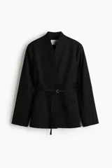 Blazer negro con cuello elevado, hombreras, abertura en los puños, bolsillos delanteros ribeteados y un cinturón fino ajustable con hebilla metálica.