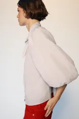 Blusa de cuello subido con escote en pico y cierre frontal con botones forrados. Presenta mangas abullonadas de largo tres cuartos con elástico en los puños y detalle de lazos en el cuello y el frente.