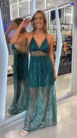 Vestido largo de fiesta color verde esmeralda, con diseño de lentejuelas brillantes en patrón lineal. Presenta un escote halter con tirantes finos, una sección central con transparencia y una falda con sobrecapa de tul transparente que revela un minivestido interior.
