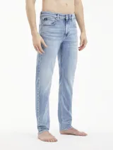 Jeans de mezclilla azul claro, tiro medio, con corte slim que se estrecha hacia el tobillo. Presentan un diseño de cinco bolsillos, bragueta con cremallera y herrajes de Calvin Klein, incluyendo una insignia de cuero en la parte trasera.