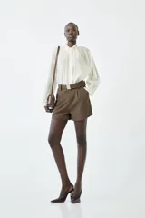 Camisa blanca de manga larga con cuello alto tipo tortuga con detalle de volados y cierre frontal abotonado. Confeccionada en tejido fluido de tencel y poliéster.